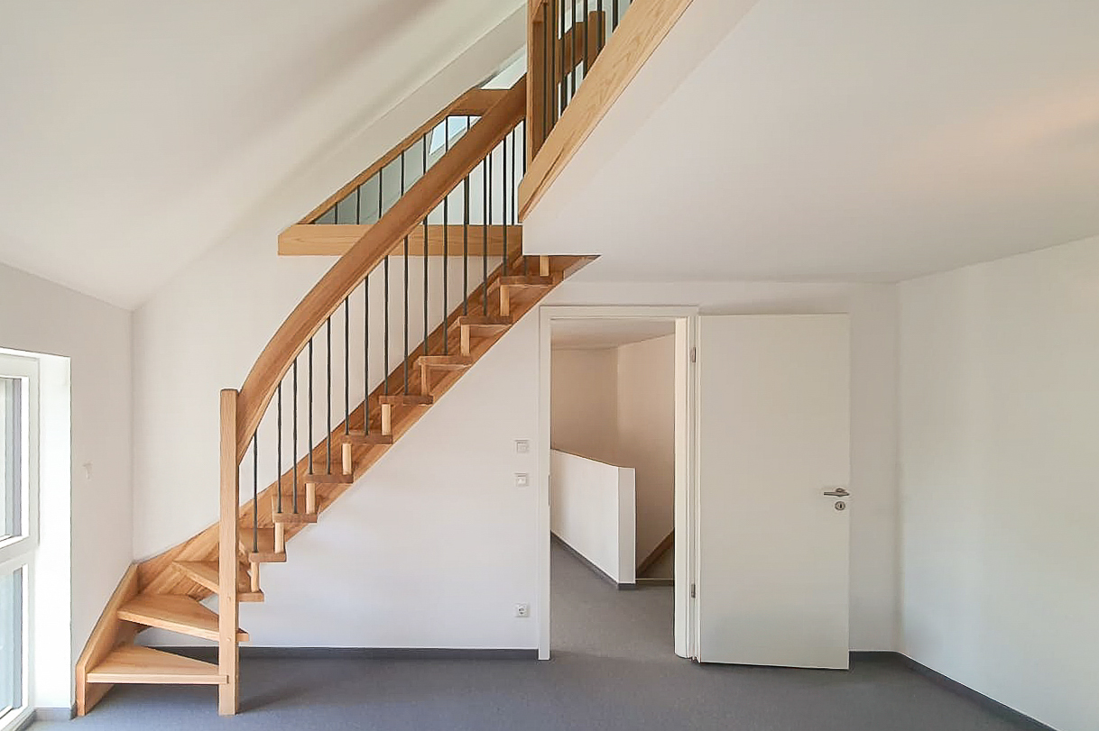 Ein moderner, heller Innenraum mit einer Holztreppe mit Metallgeländer, die zu einem oberen Dachboden führt. Der Raum hat weiße Wände, ein großes Fenster und eine offene weiße Tür.