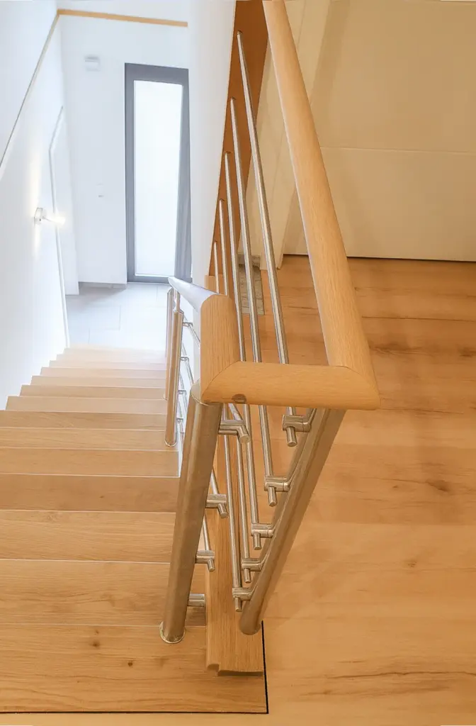 Blick von oben auf eine moderne Holztreppe mit hellem Holzgeländer und Metallgeländerstäben, die zu einem hellen Eingangsbereich mit Tür und Fliesenboden führt.