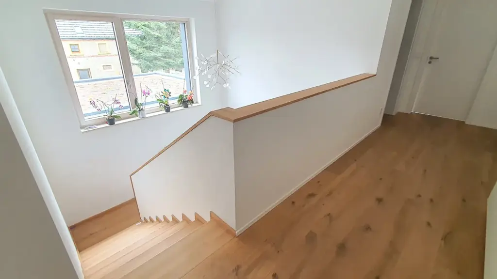 Ein heller Flur mit Holzböden, einer weißen Wand und einem Geländer entlang der Treppe sowie einem Fenster mit mehreren Topfpflanzen auf der Fensterbank, das natürliches Licht von draußen hereinlässt.