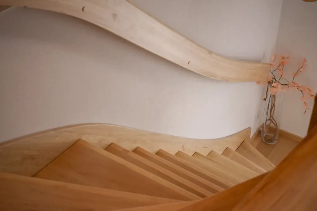 Eine moderne Holztreppe mit geschwungenen Handläufen, von oben gesehen. Die Treppe führt vor einer hellen Wand nach unten, und in der unteren Ecke steht eine dekorative Vase mit rosa Kunstblumen.