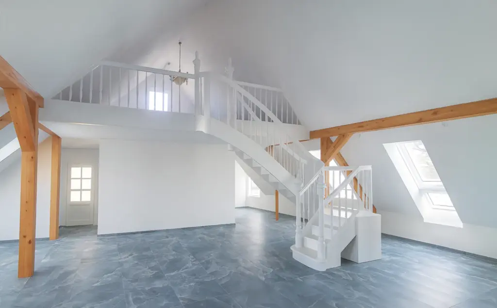 Geräumiges, helles Dachzimmer mit blau-grauem Fliesenboden, sichtbaren Holzbalken, weißen Wänden, großen Fenstern und einer weißen Treppe, die zu einem Dachboden mit Geländer führt.