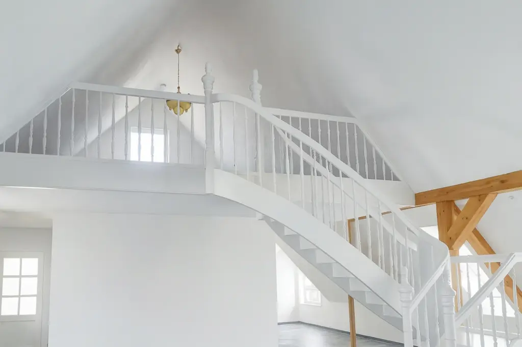 Ein helles, modernes Loft mit weißen Wänden, einer weißen Treppe mit vertikalen Geländern und Holzbalken. Durch mehrere Fenster fällt natürliches Licht in den geräumigen, offenen Innenraum.