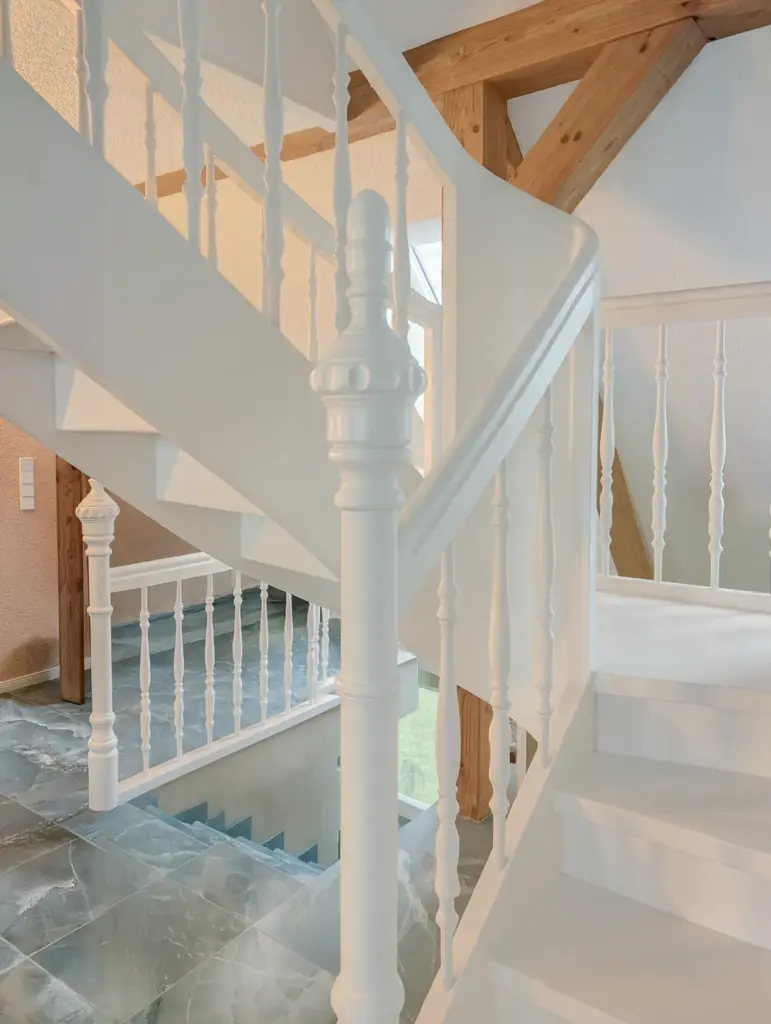 Eine weiße Holztreppe mit verschnörkelten Geländerstäben und Sprossen ist mit poliertem Marmorfußboden und sichtbaren Holzbalken an der Decke ausgestattet.