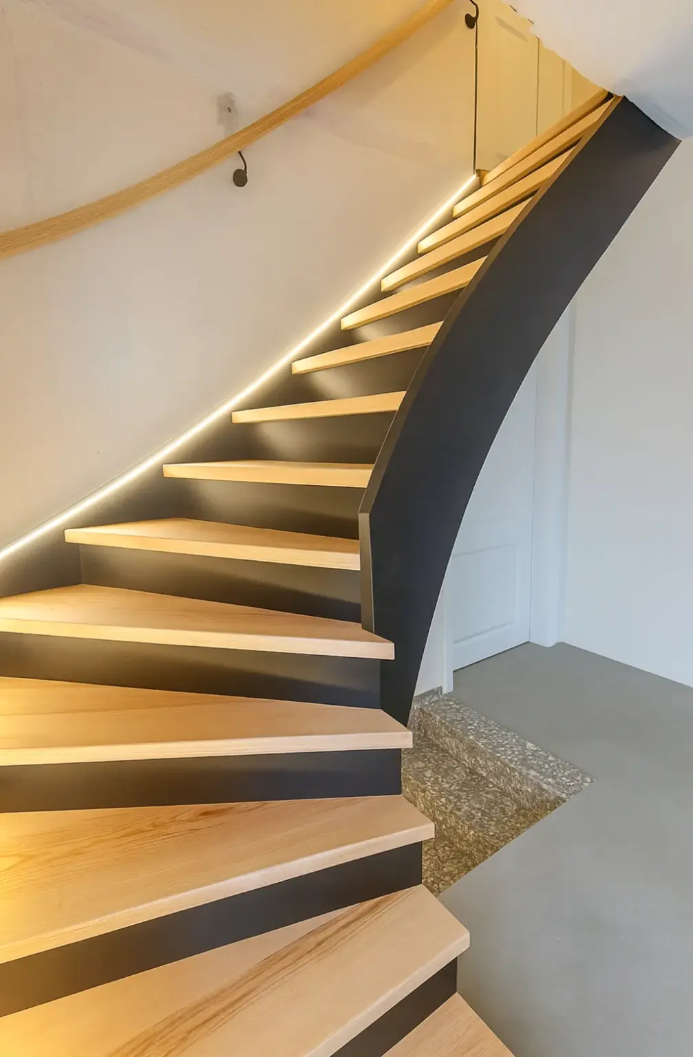 Geschwungene Holztreppe mit schwarzem Träger, beleuchtet durch eingebaute LED-Lichtbänder entlang der Wand, die zu einer weißen Tür in einem modernen Innenraum führt.