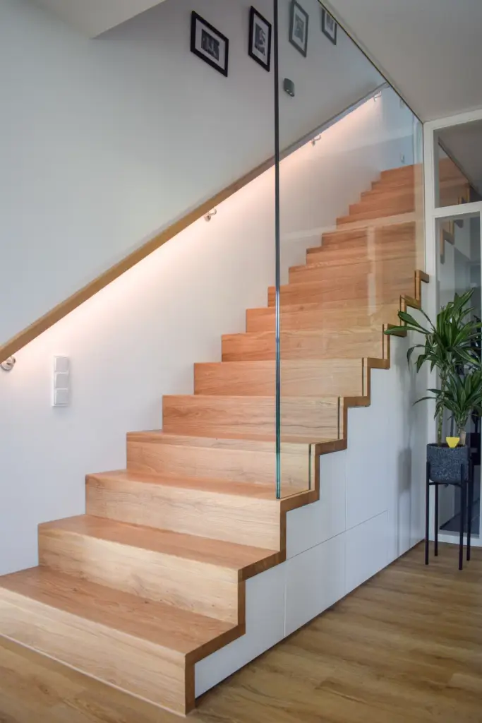 Moderne Holztreppe mit Glasgeländer, eingebauter Beleuchtung an der Wand und gerahmten Bildern darüber. Eine grüne Topfpflanze steht auf einem kleinen Tisch neben der Treppe auf einem hellen Holzboden.