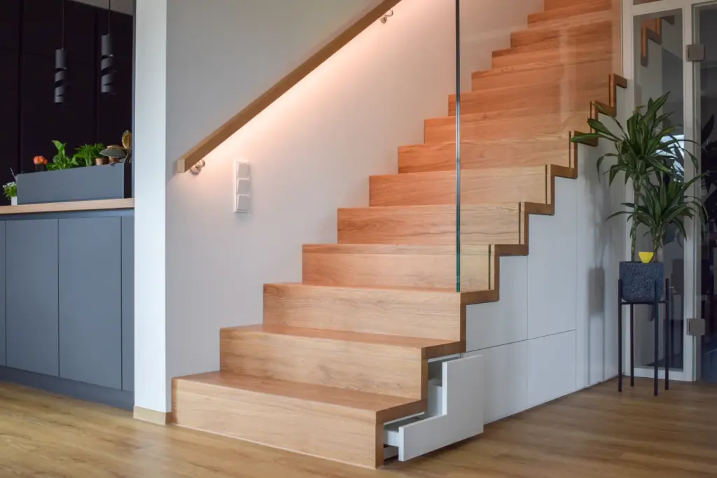 Eine moderne Holztreppe mit Glasgeländer und eingebautem Stauraum unter der Treppe, beleuchtet durch sanftes Licht, neben einer Küche mit Schränken und Topfpflanzen.