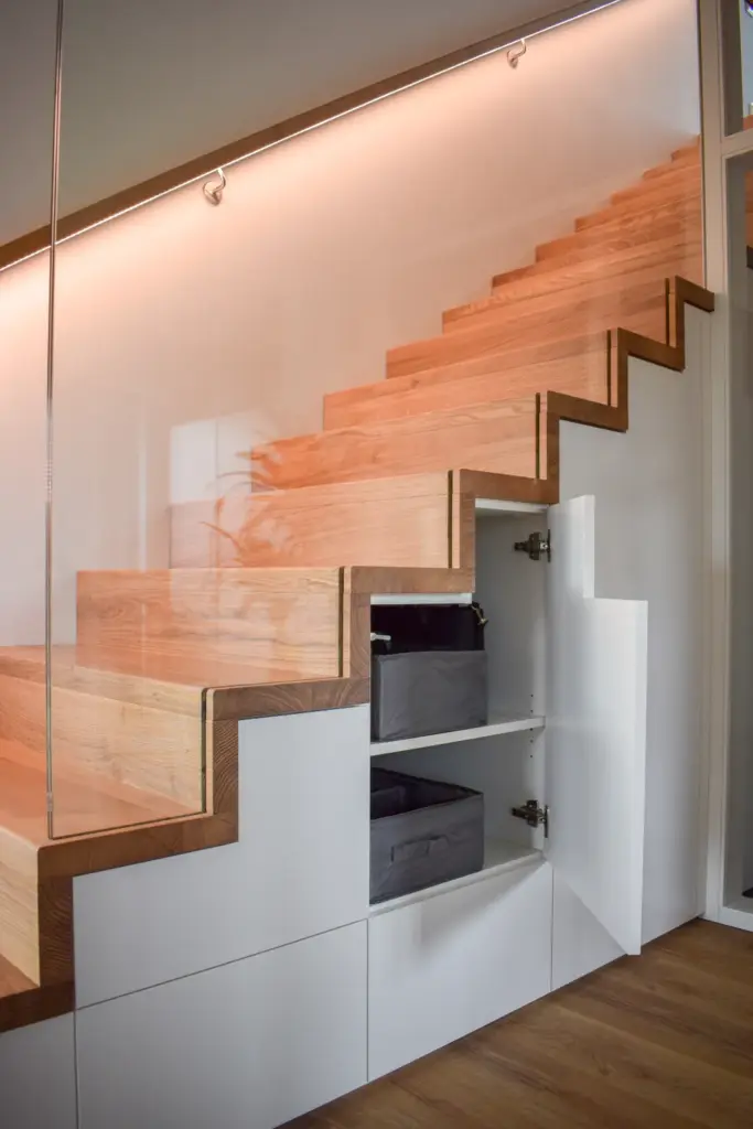 Holztreppe mit eingebauten weißen Schränken darunter, eine Tür lässt sich öffnen und gibt den Blick auf Regale mit Stoffbehältern frei. Die warme Beleuchtung unterstreicht das moderne, minimalistische Design.