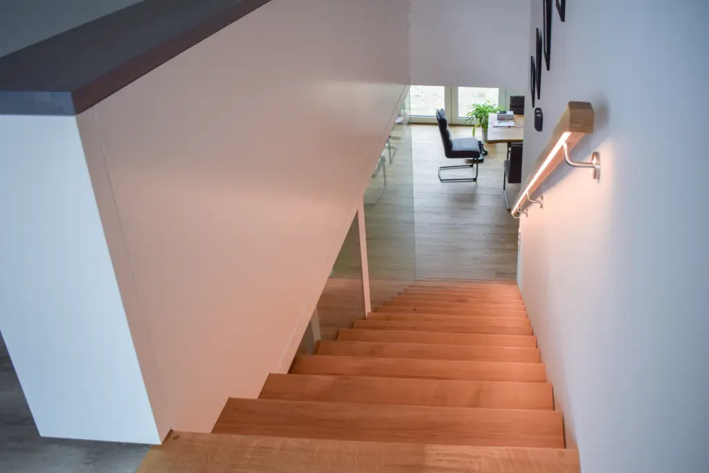 Eine moderne Holztreppe mit weißem Geländer führt hinunter in einen hellen Raum mit hellem Holzfußboden, einem Schreibtisch, einem Stuhl und einer Grünpflanze in der Nähe großer Fenster.