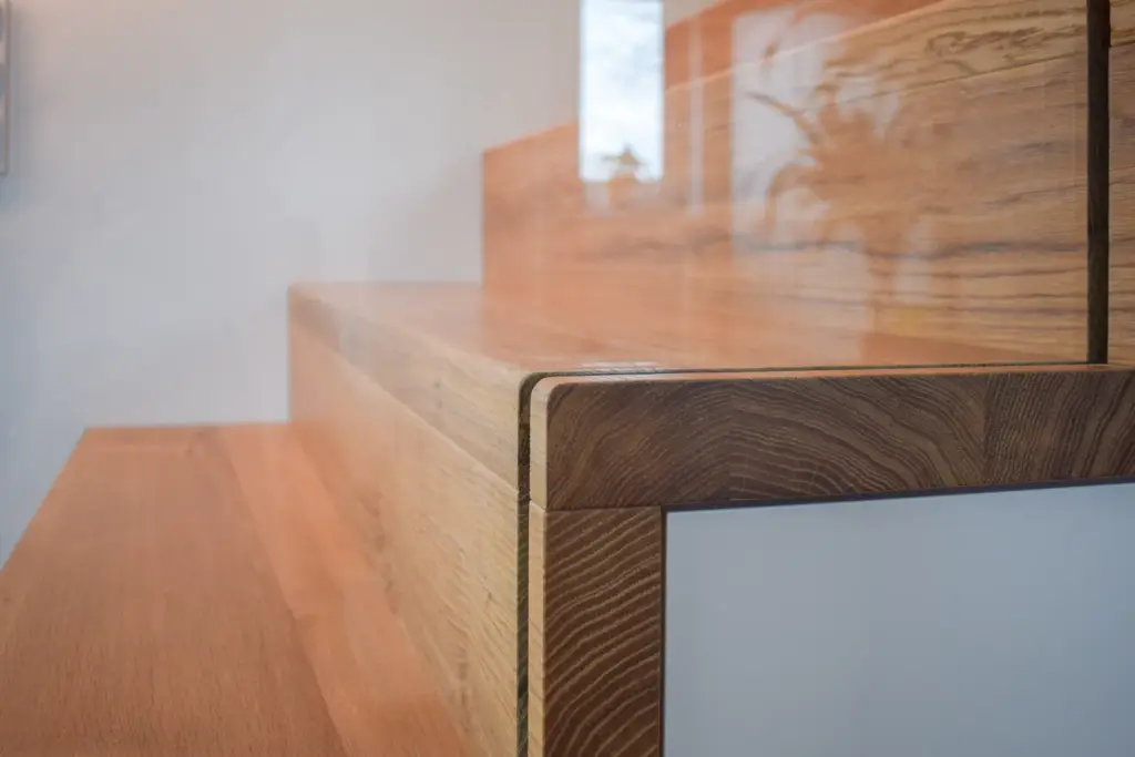 Nahaufnahme einer modernen Holztreppe mit klaren Linien und einer glatten Oberfläche, die die Maserung und Textur des Holzes zur Geltung bringt. Die Stufen haben ein minimalistisches Design und stehen vor einer hellen Wand.
