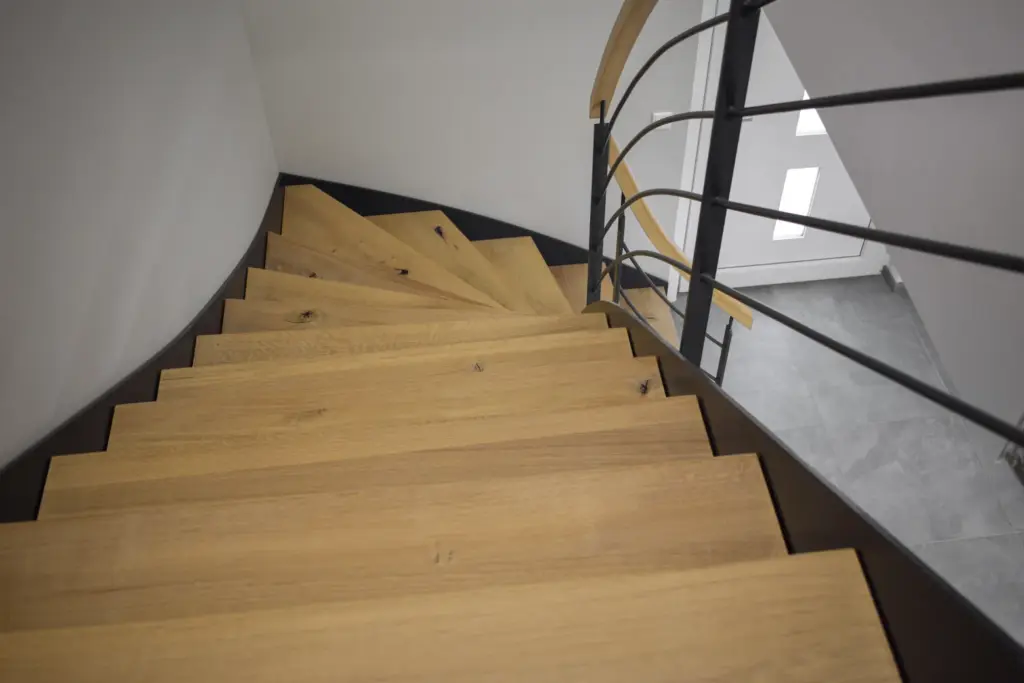 Blick von oben auf eine moderne Holztreppe mit schwarzem Metallgeländer, die geschwungen zu einem grauen Fliesenboden und einer weißen Tür in einem gut beleuchteten Innenraum hinabführt.