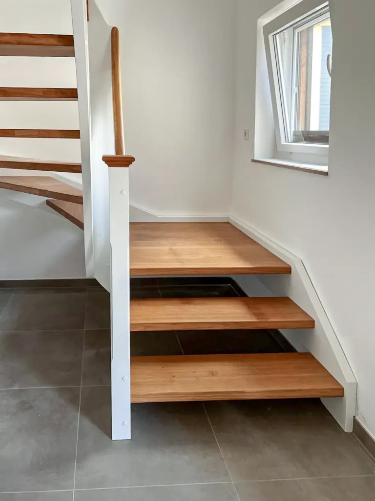 Moderne Innentreppe mit Holzstufen, weißem Geländer und einem geschwungenen Design. Der untere Teil hat drei schwimmende Stufen und grenzt an ein Fenster, das den Bereich mit natürlichem Licht erhellt. Der Boden ist in Grau gefliest.
