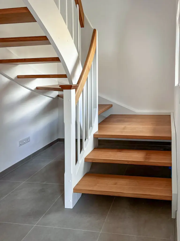 Eine moderne Holztreppe mit weißem Geländer und offenen Setzstufen in einem hellen, minimalistischen Interieur mit grauem Fliesenboden und natürlichem Licht aus einem nahe gelegenen Fenster.