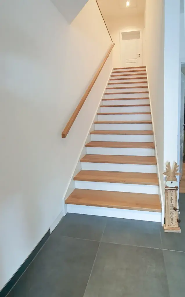 Eine Treppe mit Holzstufen und einem hölzernen Handlauf führt zu einer geschlossenen weißen Tür; ein kleines dekoratives Rentier mit Holzsockel steht rechts neben der Treppe.