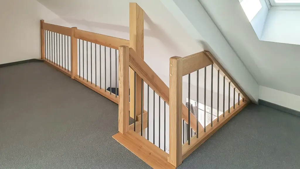 Holztreppengeländer mit vertikalen Metallstäben in einem mit Teppichboden ausgelegten, loftartigen Innenraum mit schräger Decke und Oberlichtfenster, das für natürliches Licht sorgt.