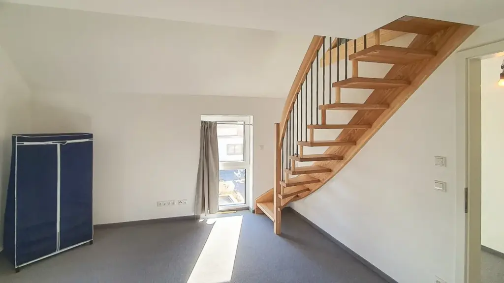 Ein heller, minimalistischer Raum mit grauem Teppich, einer hölzernen Wendeltreppe, einem blauen Stoffschrank und einem Fenster mit einem einzigen Vorhang, der das Sonnenlicht hereinlässt.