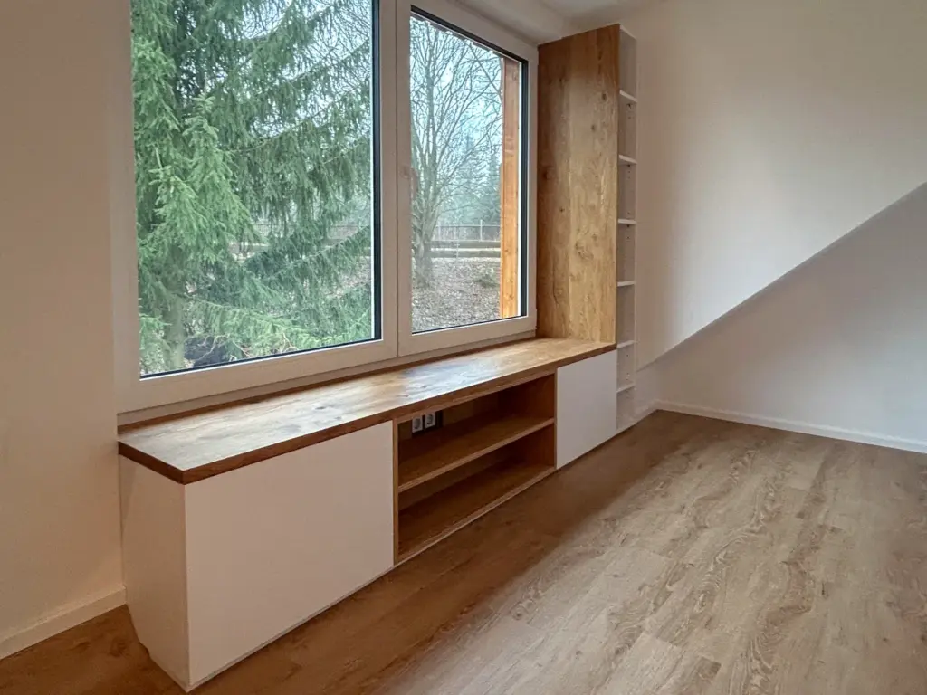 Eine eingebaute Fensterbank aus Holz mit weißen Schränken und offenen Regalen steht neben großen Fenstern mit Blick auf Bäume in einem hellen, minimalistischen Raum mit hellem Holzfußboden.