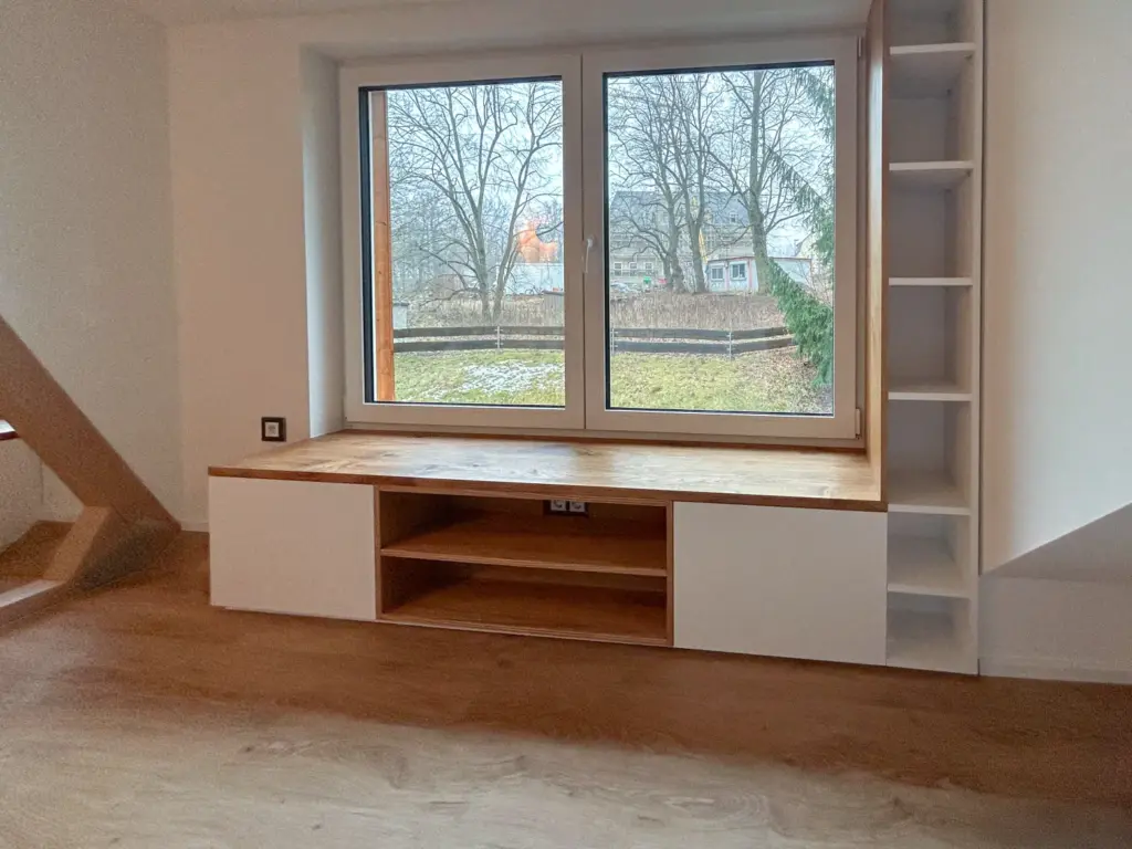 Eine große Fensterbank mit hölzerner Oberfläche, flankiert von Regalen auf der rechten Seite und Ablagefächern darunter, bietet einen Blick auf einen winterlichen Garten mit kahlen Bäumen.