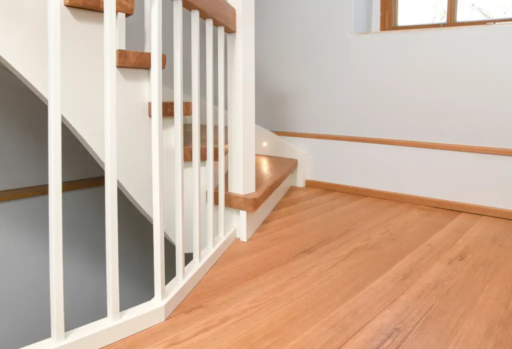 Eine moderne Innentreppe mit Holzstufen und weißem Geländer neben einem hellen Holzboden und hellgrauen Wänden. An der Seite der Treppe ist eine kleine Einbaulampe eingebaut.