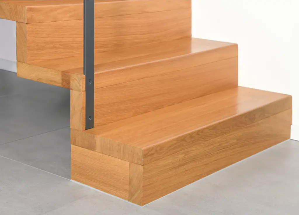 Nahaufnahme von drei Holztreppenstufen mit glatter Oberfläche, einem Metallhandlauf auf der linken Seite und hellgrauen Bodenfliesen sowie einer weißen Wand im Hintergrund.