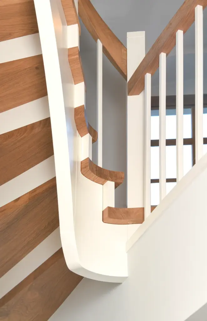 Nahaufnahme einer modernen Treppe mit weißem Geländer und hellen Holzstufen, mit glatten, geschwungenen Designdetails und vertikalen Geländerstäben, vor einer neutralgrauen Wand und einem Fenster im Hintergrund.