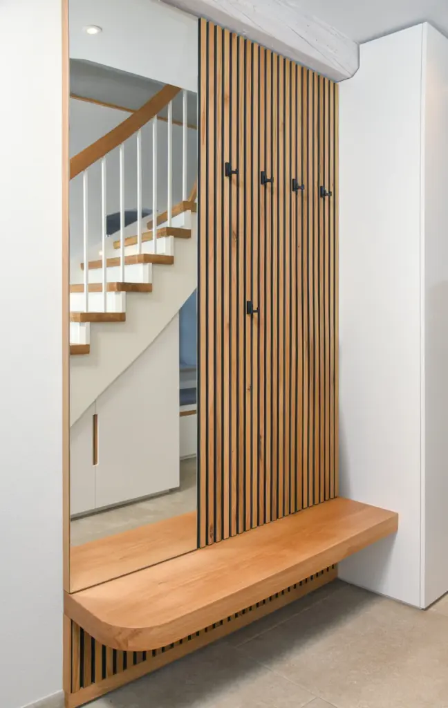 Ein moderner Eingangsbereich mit einem Wandpaneel aus Holzlatten mit Haken, einer Holzbank darunter, einem Spiegel und einer Treppe mit weißem Geländer im Hintergrund. Das Design ist minimalistisch und klar.