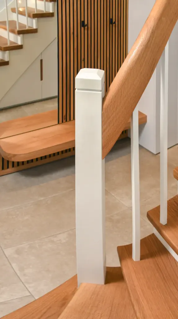 Nahaufnahme einer modernen Holztreppe mit hellbraunen Stufen, weißen Geländern und einem quadratischen weißen Pfosten vor einem gefliesten Boden und einem holzgetäfelten Hintergrund.