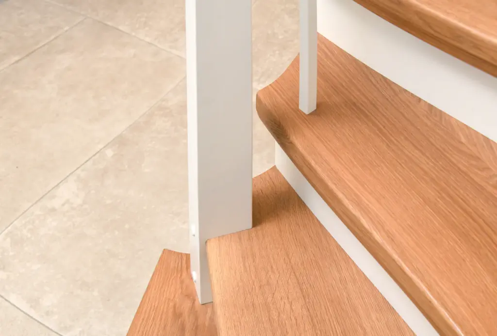 Nahaufnahme von Holztreppenstufen mit weißen Setzstufen und einer weißen vertikalen Stütze vor einem beigefarbenen Fliesenboden.