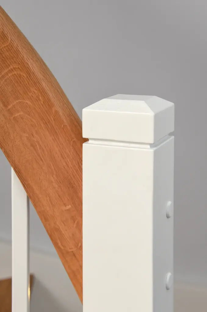 Nahaufnahme eines weißen, quadratischen Treppenpfostens mit einer dekorativen Kappe neben einem Holzhandlauf mit natürlicher Oberfläche vor einem neutralgrauen Hintergrund.