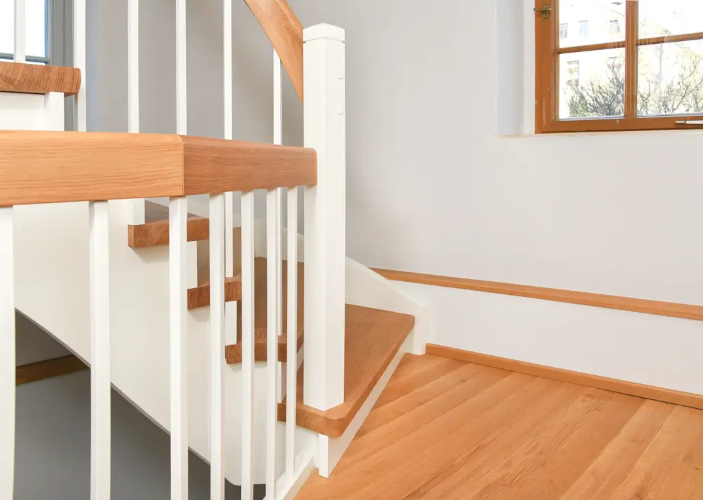 Eine moderne Holztreppe mit weißem Geländer und hellbraunen Stufen, neben einem Fenster, das natürliches Licht hereinlässt, in einem hellen, sauberen Raum mit Holzfußboden.