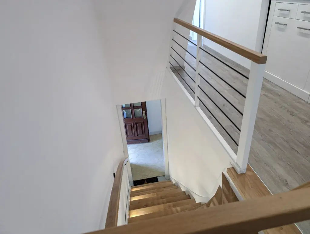 Eine Holztreppe mit weißem Metallgeländer führt hinunter zu einem gefliesten Eingangsbereich mit einer braunen Tür. Das Treppenhaus hat weiße Wände, und im Obergeschoss sind Holzfußböden und ein Teil einer Küche zu sehen.