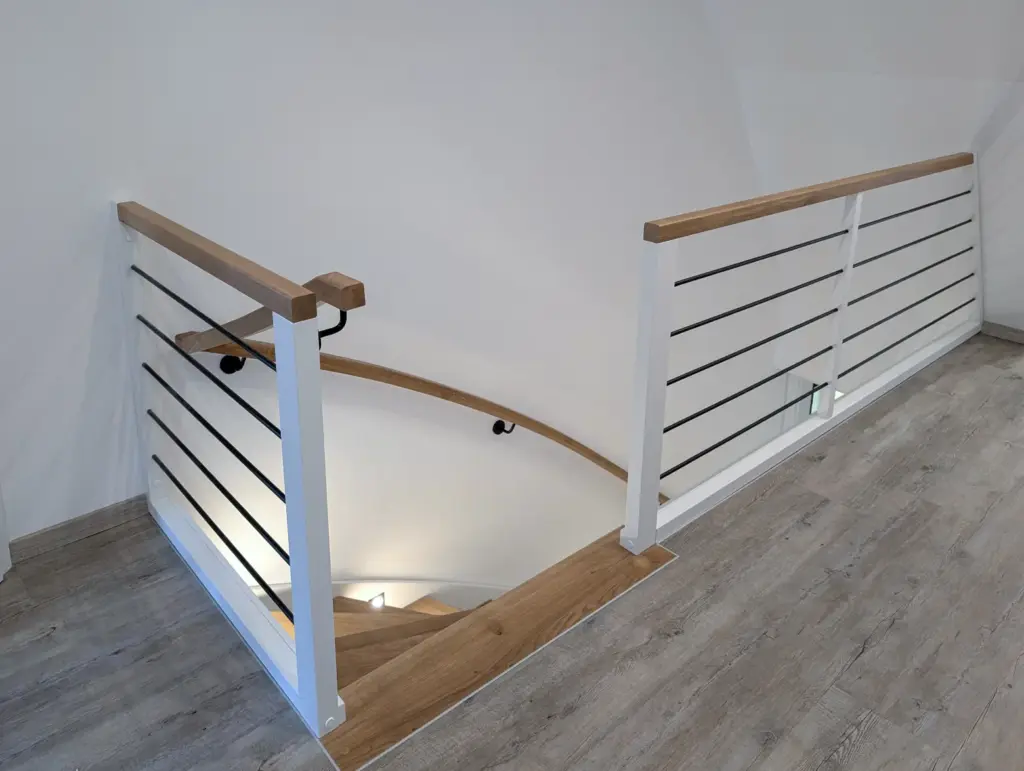 Eine moderne Innentreppe mit Holzhandläufen, weißen Pfosten und schwarzen horizontalen Metallstäben, die von einem Raum mit hellem Holzfußboden und weißen Wänden hinunterführt.