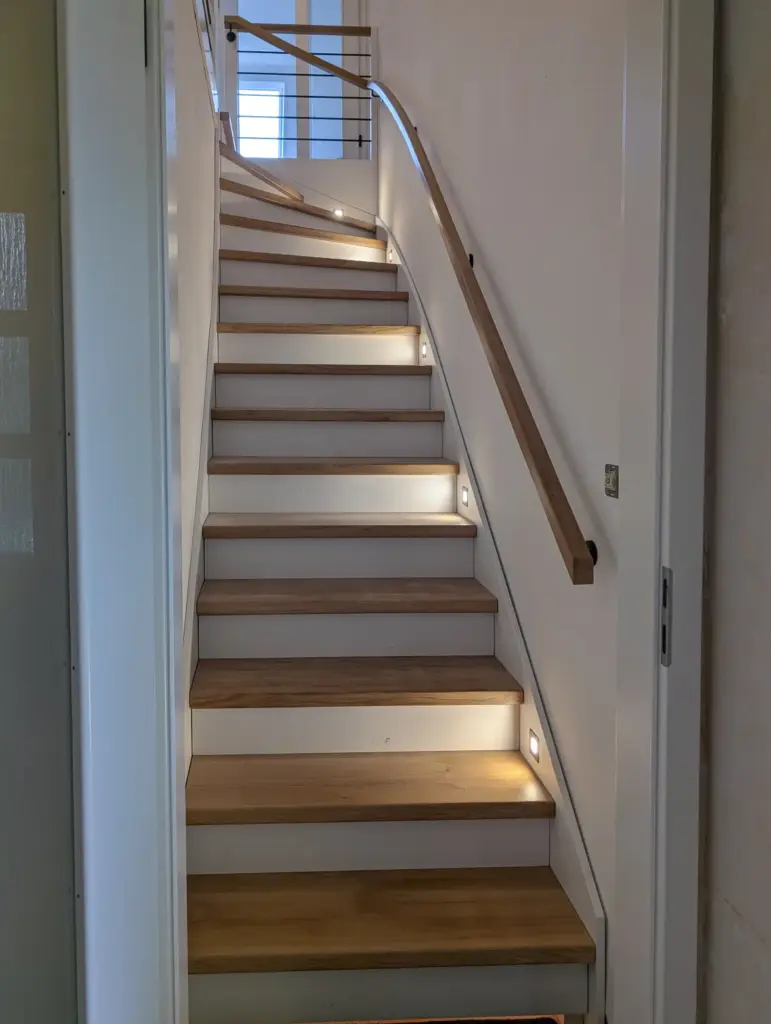 Eine moderne Holztreppe mit eingebauter Stufenbeleuchtung und einem Holzhandlauf auf der rechten Seite führt zu einem hellen, fensterbeleuchteten Treppenabsatz.