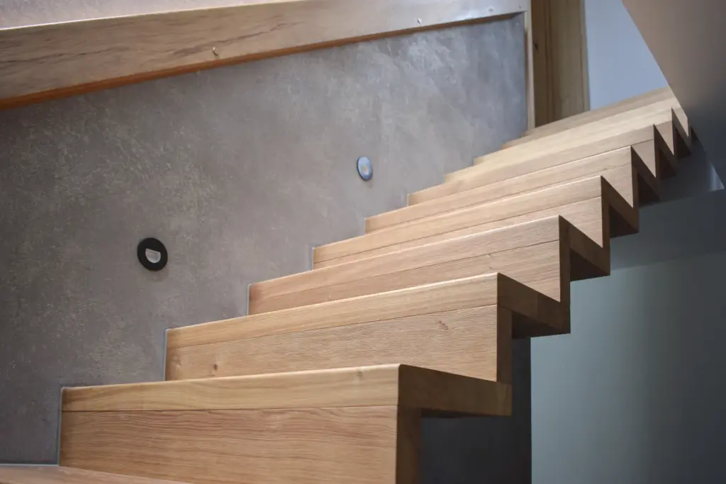 Eine moderne Holztreppe mit Zickzackmuster, angrenzend an eine glatte graue Wand mit zwei kleinen runden schwarzen Leuchten. Natürliches Licht beleuchtet die Treppe und unterstreicht ihre klaren, kantigen Linien.