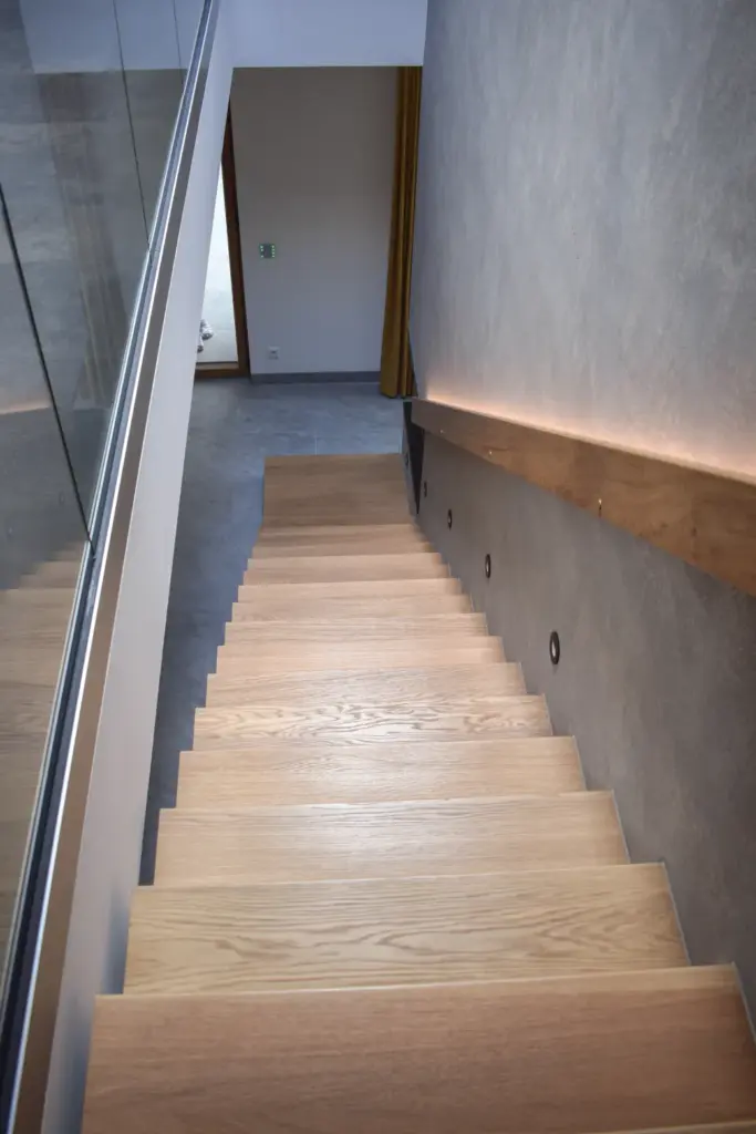 Eine moderne Holztreppe mit hellen Stufen führt nach unten, begrenzt von einem Glasgeländer auf der linken Seite und einer grauen Wand mit runden Leuchten und einem Holzhandlauf auf der rechten Seite.