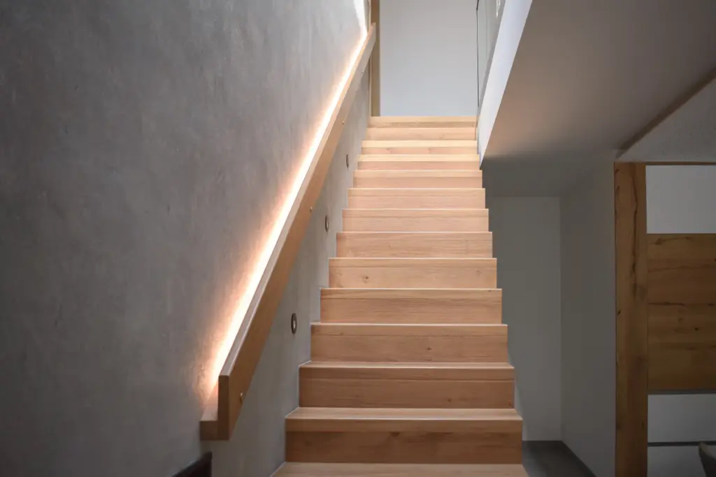 Eine moderne Holztreppe mit eingebauter Seitenbeleuchtung führt neben einer grauen Wand in einem minimalistischen Innenraum nach oben. Die Treppe hat klare Linien und einen einfachen Handlauf.