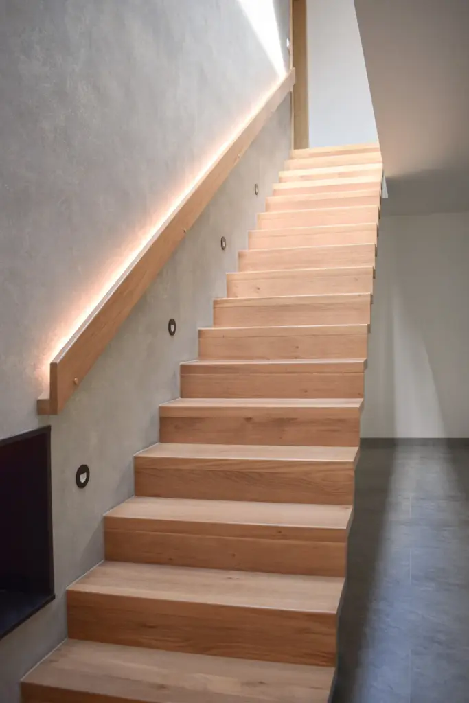 Eine moderne Holztreppe ohne sichtbare Stützen führt neben einer hellgrauen Wand mit eingebauter Handlaufbeleuchtung nach oben und sorgt für ein minimalistisches und modernes Aussehen.