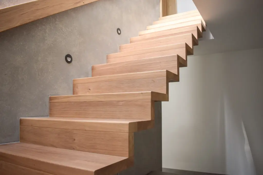 Schwebende Holztreppe mit dicken, rechteckigen Stufen, die an einer Betonwand befestigt ist, mit minimalistischen runden Wandleuchten und einem modernen, offenen Design. Das Licht scheint aus dem oberen Stockwerk.