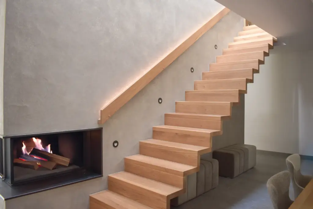 Modernes Interieur mit schwebender Holztreppe, minimalistischen grauen Wänden, eingebauter LED-Handlaufbeleuchtung und einem Kamin mit brennendem Feuer unter der Treppe. Unter der Treppe sind Würfelhocker versteckt.