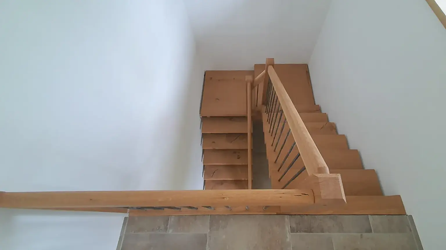Blick auf eine Holztreppe mit Geländer, die abrupt an einer Wand endet, wodurch die Treppe unbenutzbar wird. Die umgebenden Wände sind weiß, und der Boden am oberen Ende der Treppe ist mit großen beigen Fliesen ausgelegt.