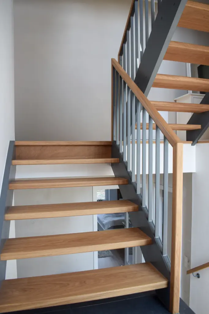 Eine moderne Treppe mit Holzstufen und grauem Geländer, die durch ihre offene Gestaltung viel Licht durchlässt und in einem hellen Innenraum in ein höheres Stockwerk führt.