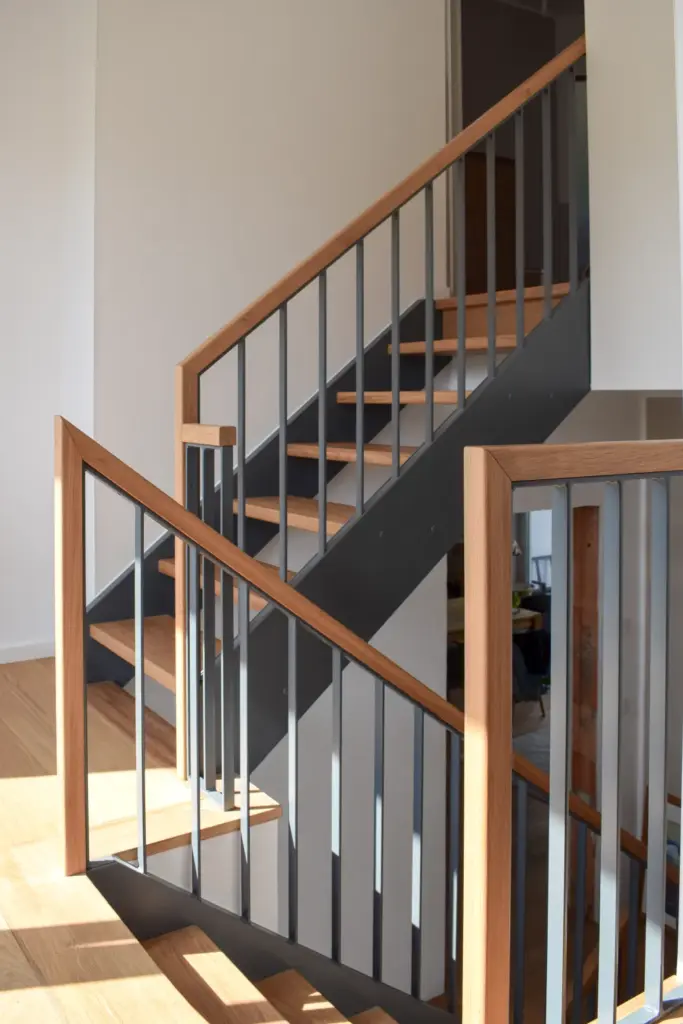 Eine moderne Treppe mit schwarzen Metallstäben und Handläufen aus hellem Holz, beleuchtet von natürlichem Sonnenlicht, vor weißen Wänden und einem Holzboden.