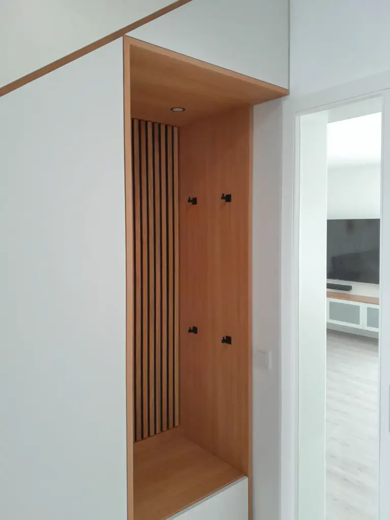 Eine moderne Holznische mit vertikalen Latten, schwarzen Kleiderhaken und einer Bank darunter, eingebaut in eine weiße Wand neben einer Glastür in einem hellen, minimalistischen Raum.