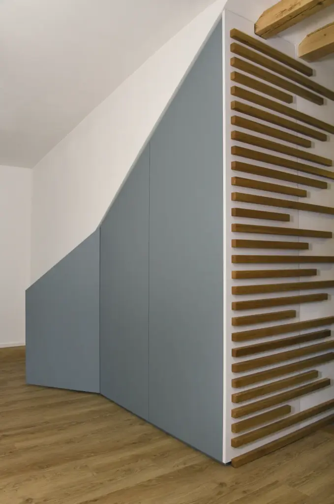 Moderner Stauraum unter der Treppe mit eleganten blaugrauen Schranktüren und einem vertikalen Holzlattenmuster auf der rechten Seite, vor einer weißen Wand und einem Holzboden.