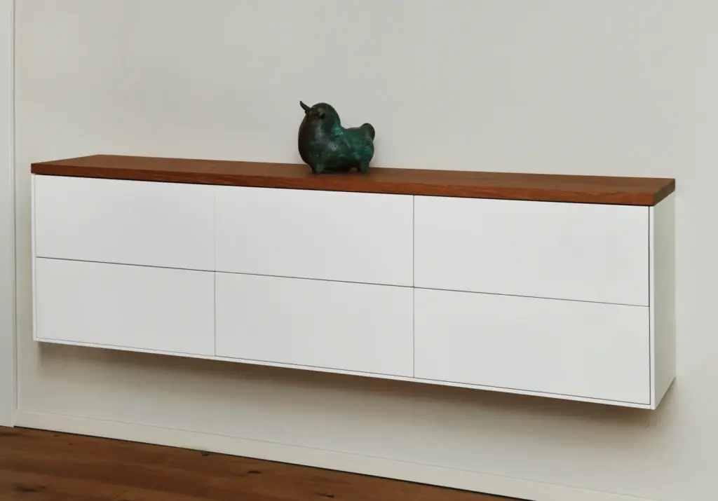 Ein minimalistischer weißer Wandschrank mit sechs Schubladen, die mit einer hölzernen Oberfläche versehen sind, und einer kleinen dekorativen Vogelskulptur auf der Oberseite. Der Schrank steht vor einer schlichten, hellen Wand.