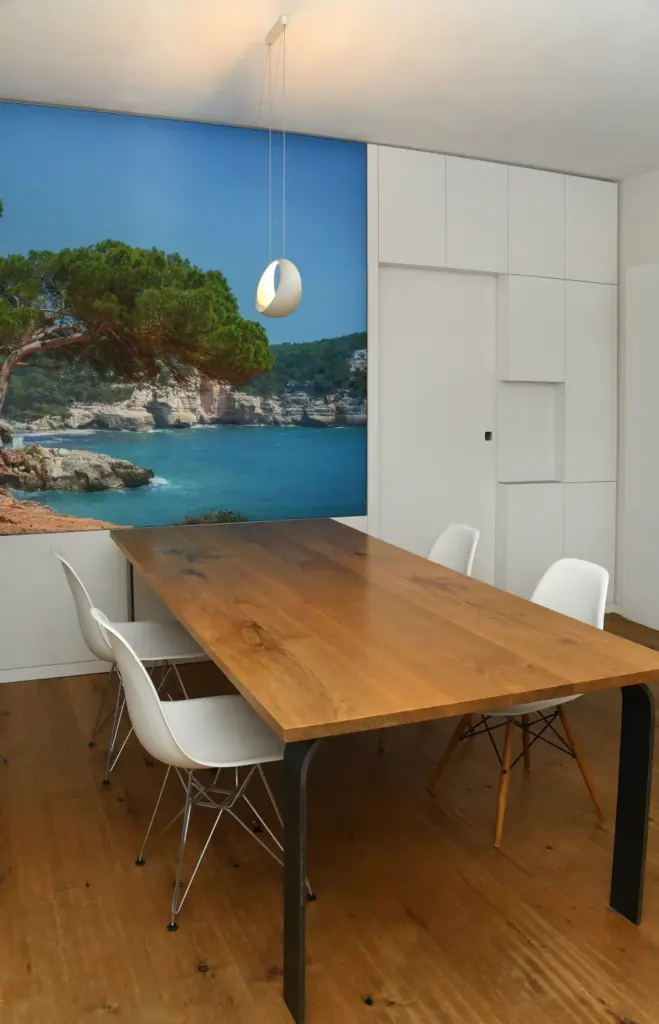 Modernes Esszimmer mit einem Holztisch, vier weißen Stühlen und einer Wandmalerei, die eine Küstenszene mit blauem Wasser, Klippen und Bäumen zeigt; im Hintergrund sind weiße Schränke zu sehen.