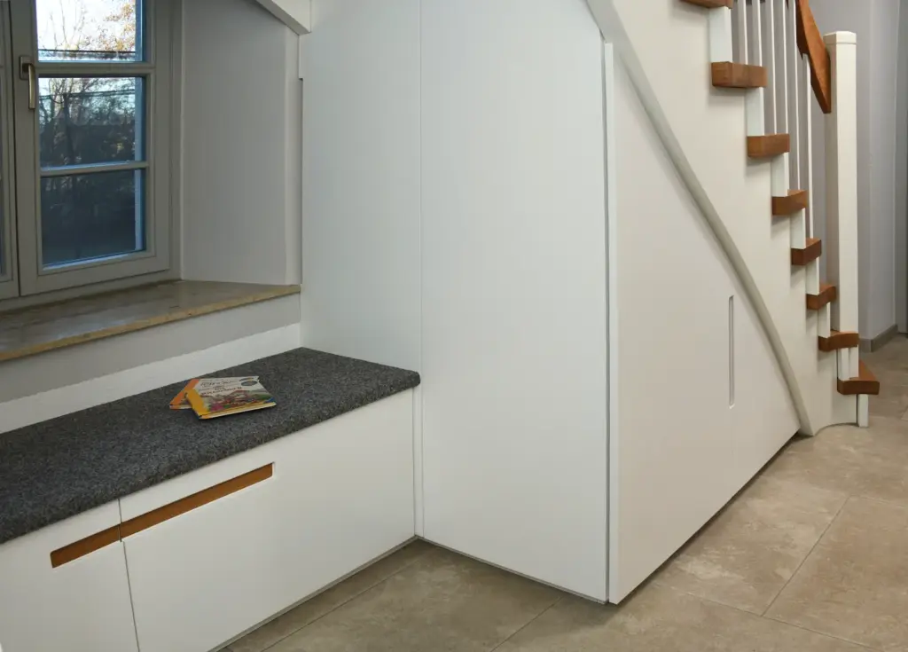 Ein moderner Flur mit weißen Einbauschränken unter einer Treppe mit Holzstufen und Handlauf. Unter einem Fenster steht eine graue Steinbank, auf der eine Zeitschrift liegt.