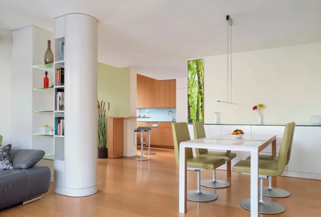 Moderner offener Küchen- und Essbereich mit hellem Holzfußboden, weißen Wänden, grünen Esszimmerstühlen, einem weißen Tisch, eingebauten Regalen, einer Säule und großen Fenstern, die viel Tageslicht hereinlassen.