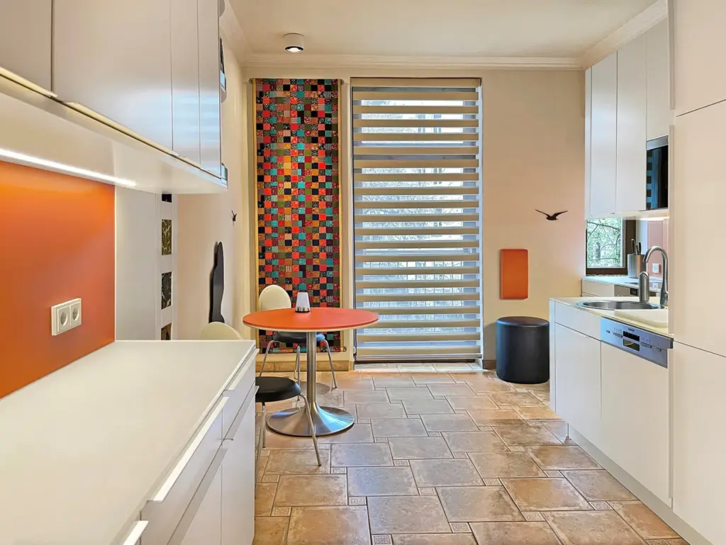 Moderne Küche mit weißen Schränken, orangefarbener Aufkantung und einem kleinen runden orangefarbenen Tisch mit zwei Stühlen. Im Hintergrund befinden sich ein Fenster mit gestreiften Jalousien und eine bunte Mosaikwand. Der Boden ist in einem warmen Ton gefliest.