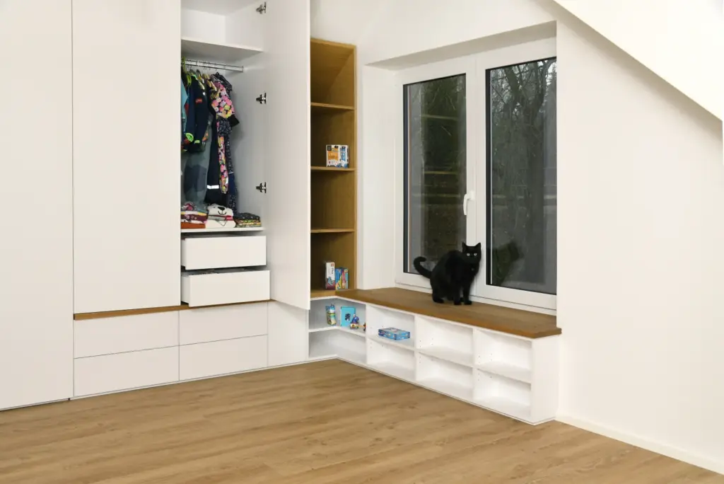 Eine schwarze Katze sitzt auf einer Holzbank am Fenster in einem hellen, modernen Raum mit weißen Schränken, Regalen, einem offenen Kleiderschrank und einem hellen Holzboden.