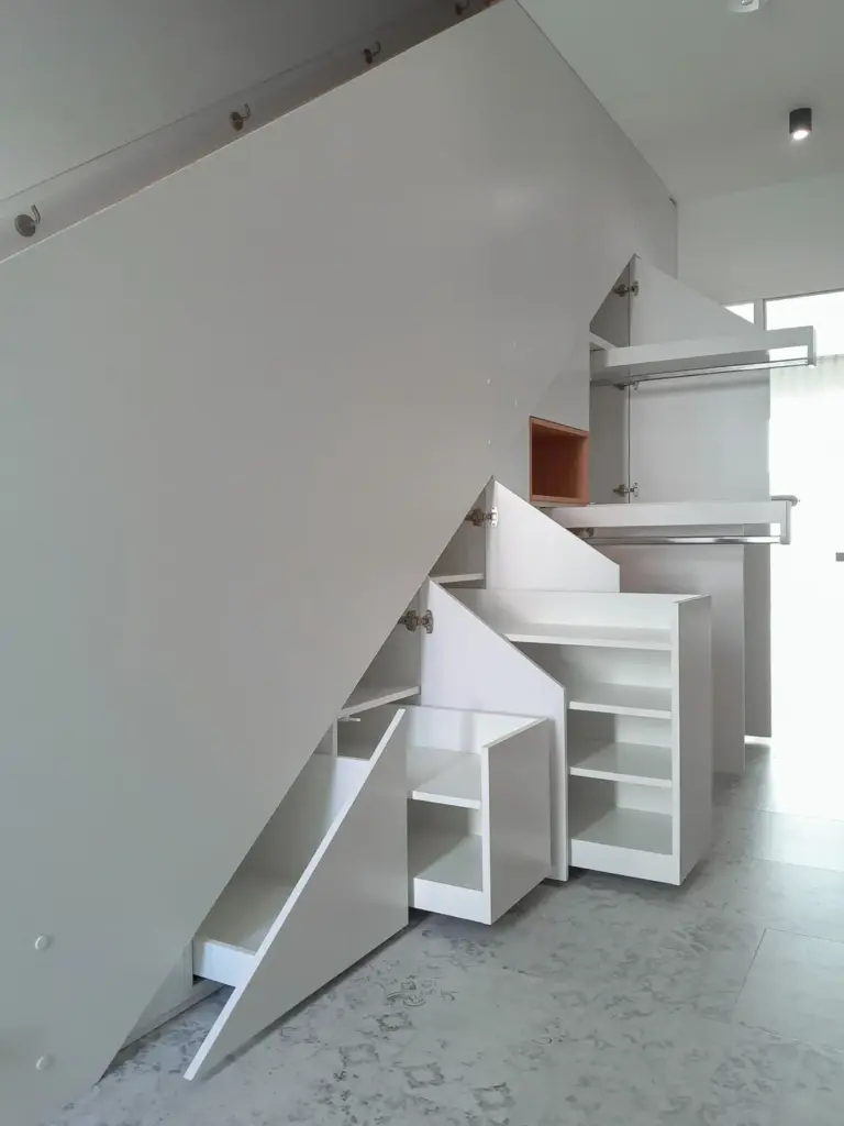 Weiße Unterflurschränke mit mehreren Einlegeböden und ausziehbaren Fächern, die den Platz unter einer Treppe in einem modernen, minimalistischen Interieur maximieren.